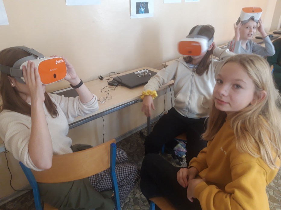 Grupa 8 uczniów siedzi przy stołach w klasie. Kilkoro uczniów trzyma w rękach urządzenie: okulary VR, kilkoro innych ma okulary VR założone na głowę. 