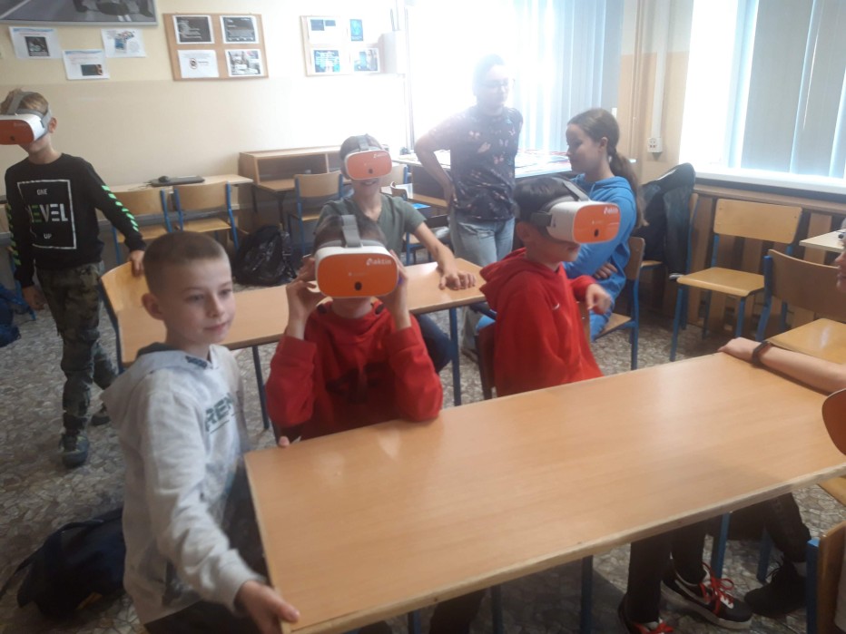 Grupa 8 uczniów siedzi przy stołach w klasie. Kilkoro uczniów trzyma w rękach urządzenie: okulary VR, kilkoro innych ma okulary VR założone na głowę. 